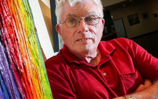 Julian is wearing a red polo shirt and standing in front of a painting featuring heavy rainbow coloured oil paint. He has white hair and blue eyes and he's wearing glasses. His expression is neutral