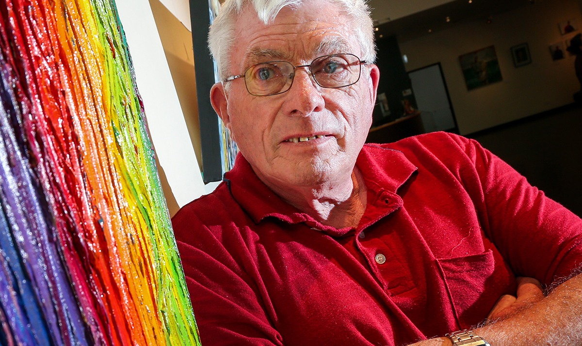 Julian is wearing a red polo shirt and standing in front of a painting featuring heavy rainbow coloured oil paint. He has white hair and blue eyes and he's wearing glasses. His expression is neutral